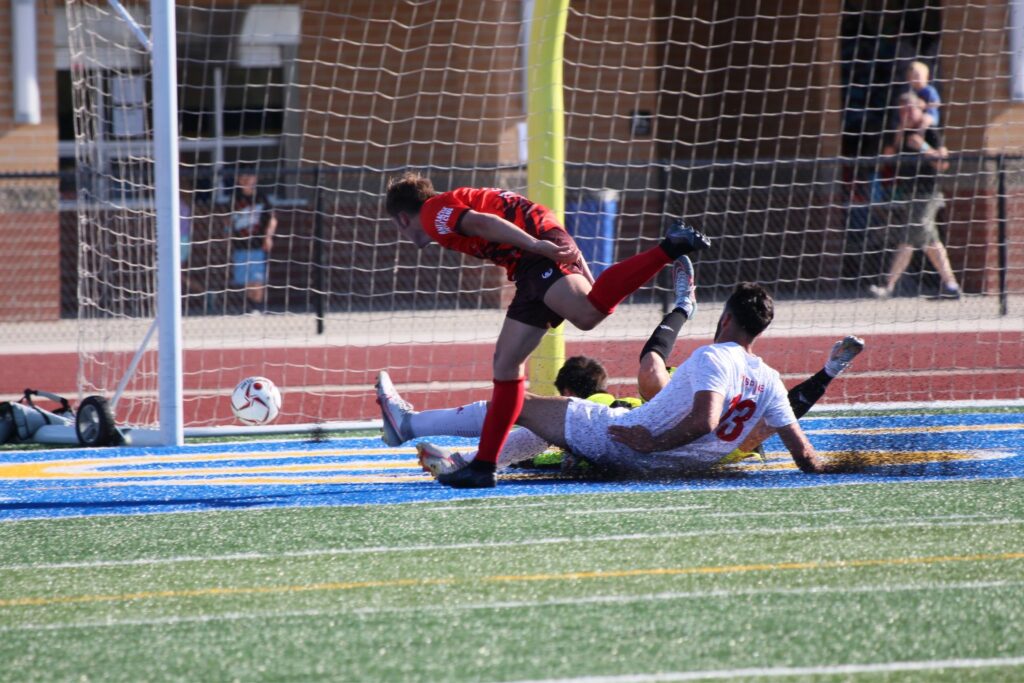 Photo of Jack Voight scoring for Lansing Common FC as a Cedars FC defender slides and the goalkeeper dives to attempt to save the ball. 
